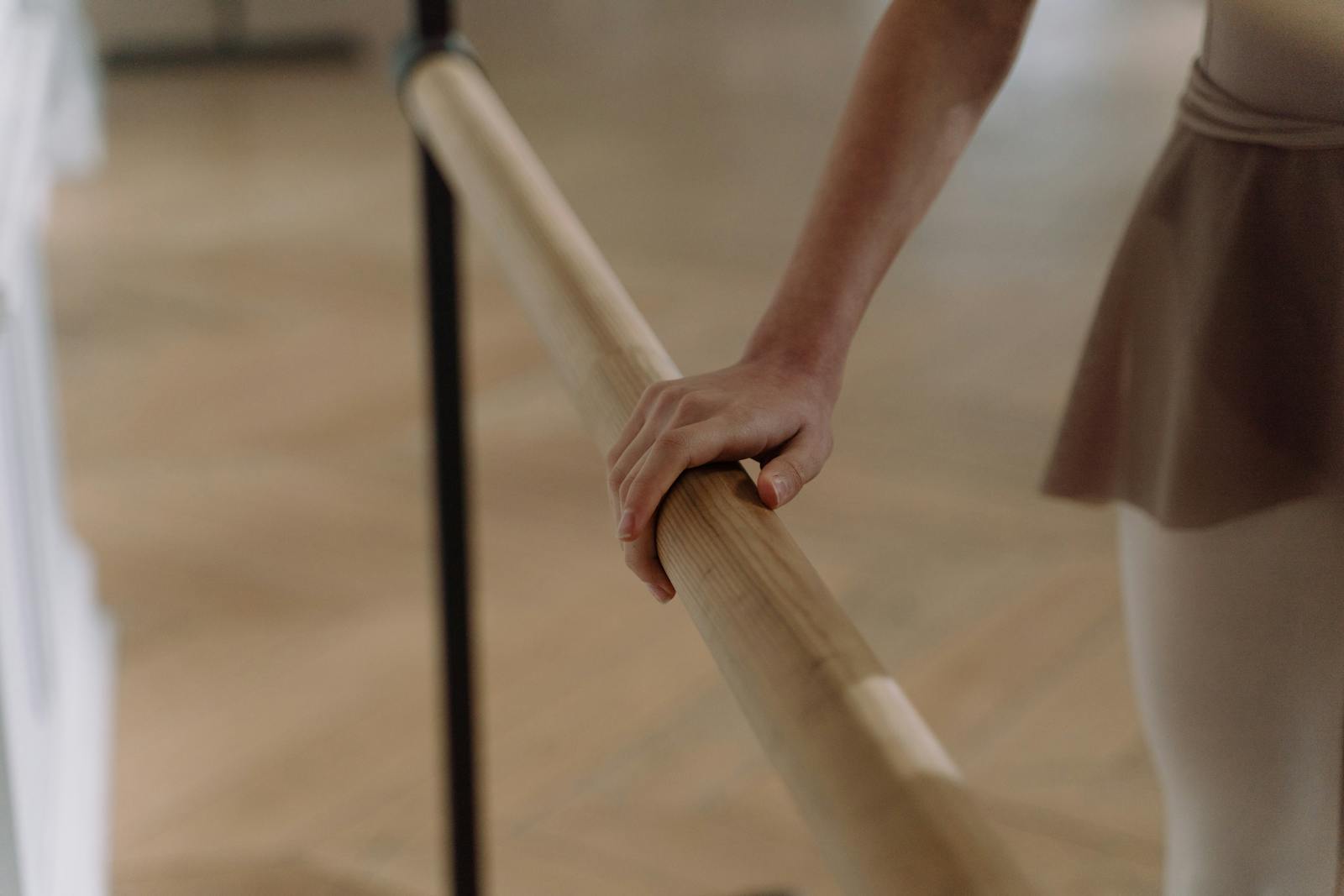 A dancer's hand resting on the wooden barre — the intimate detail of class work, parquet floor in soft focus