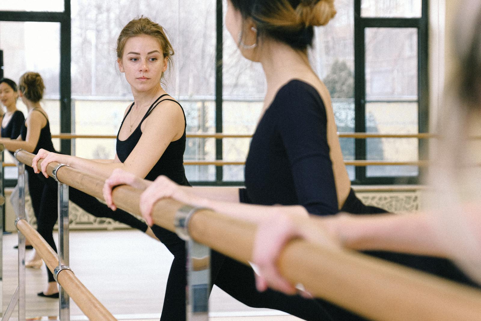 Adult students in black leotards at the wooden barre in a classical ballet studio — focused practice, natural daylight
