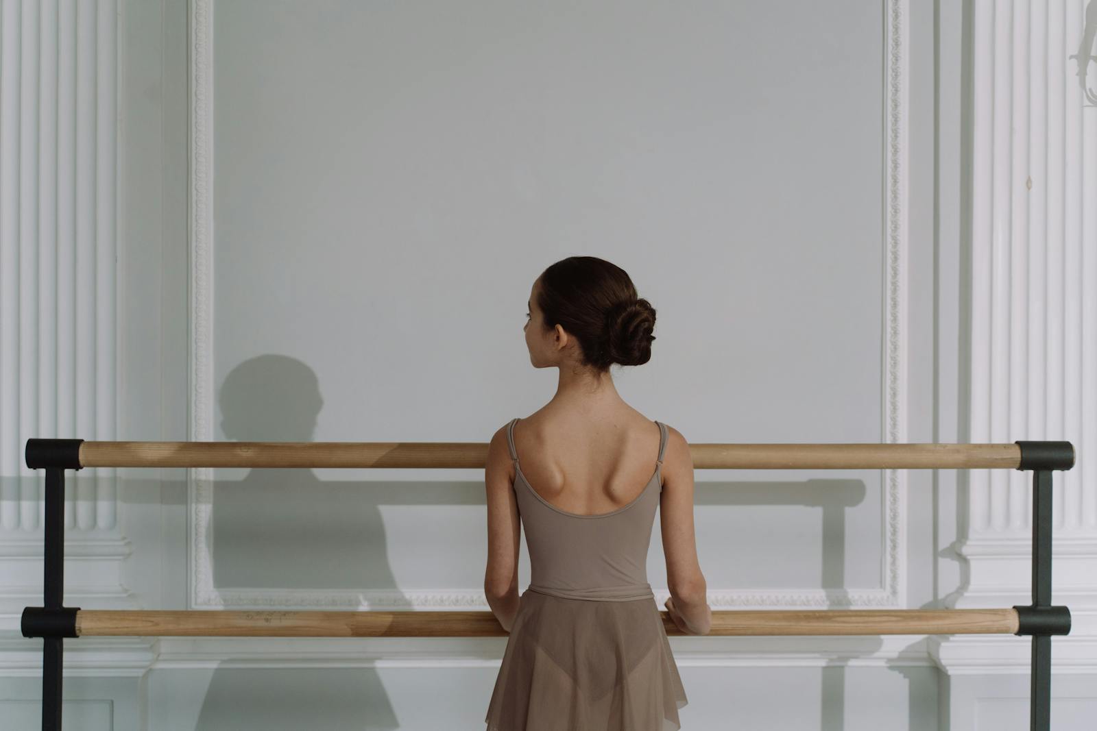Back view of an adult dancer at the double barre in a classical paneled studio — the spine as the subject