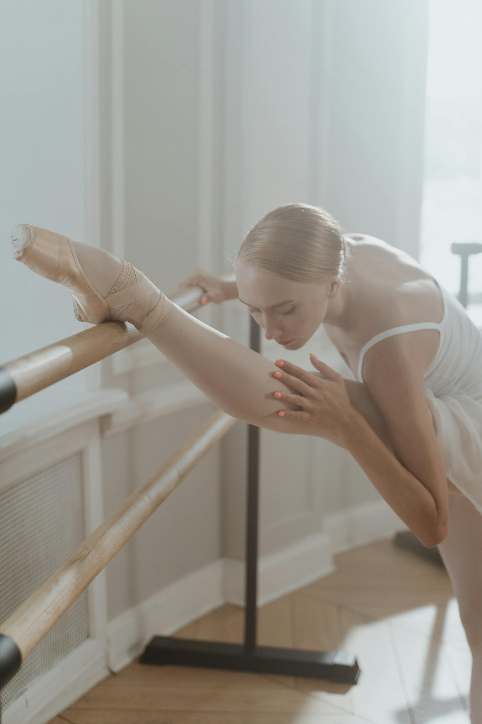 Adult dancer stretching at the barre in a sunlit classical studio — measured, careful practice