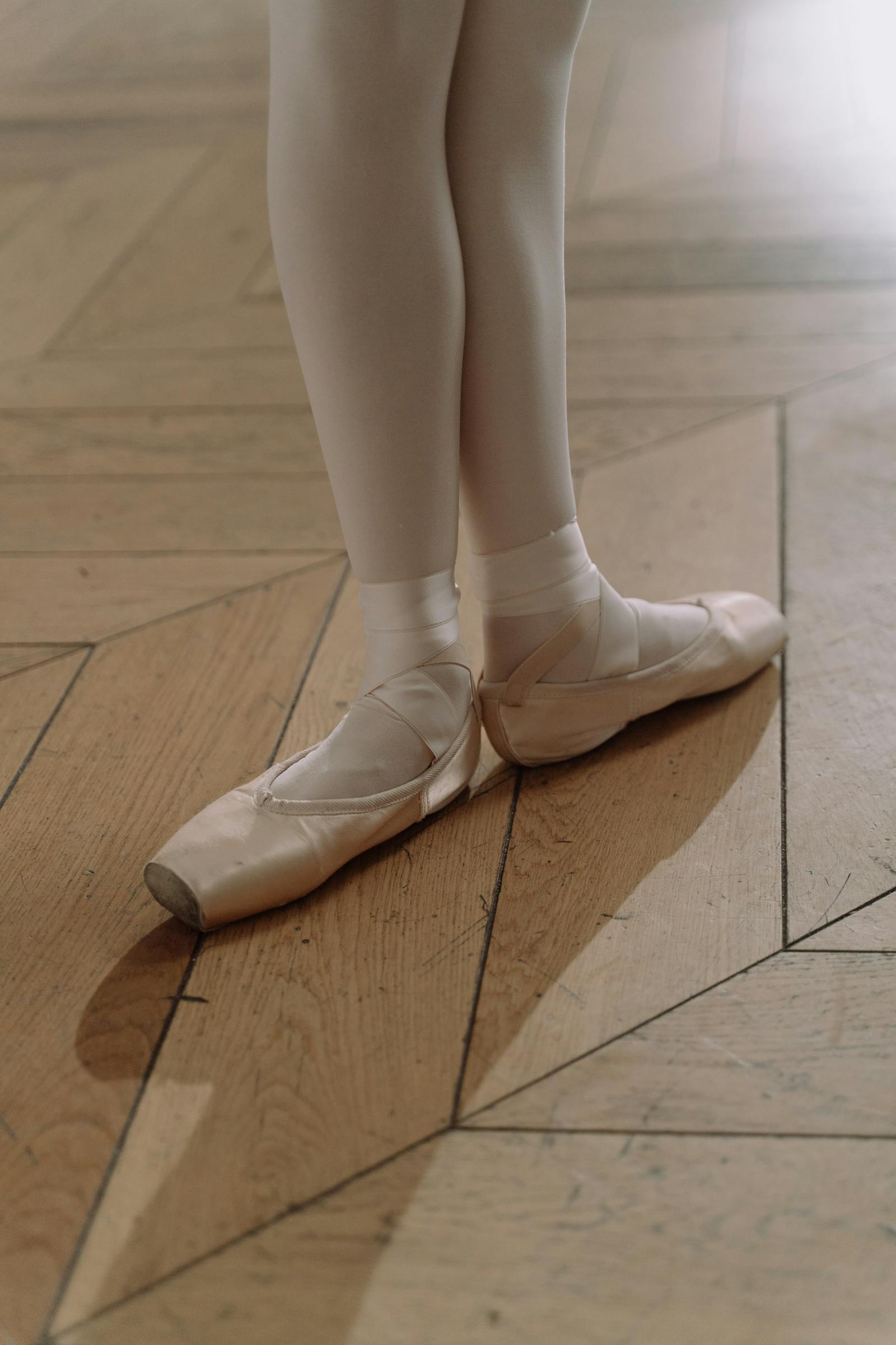 Adult dancer's feet in pointe shoes on a herringbone parquet floor — the work of years, not weeks