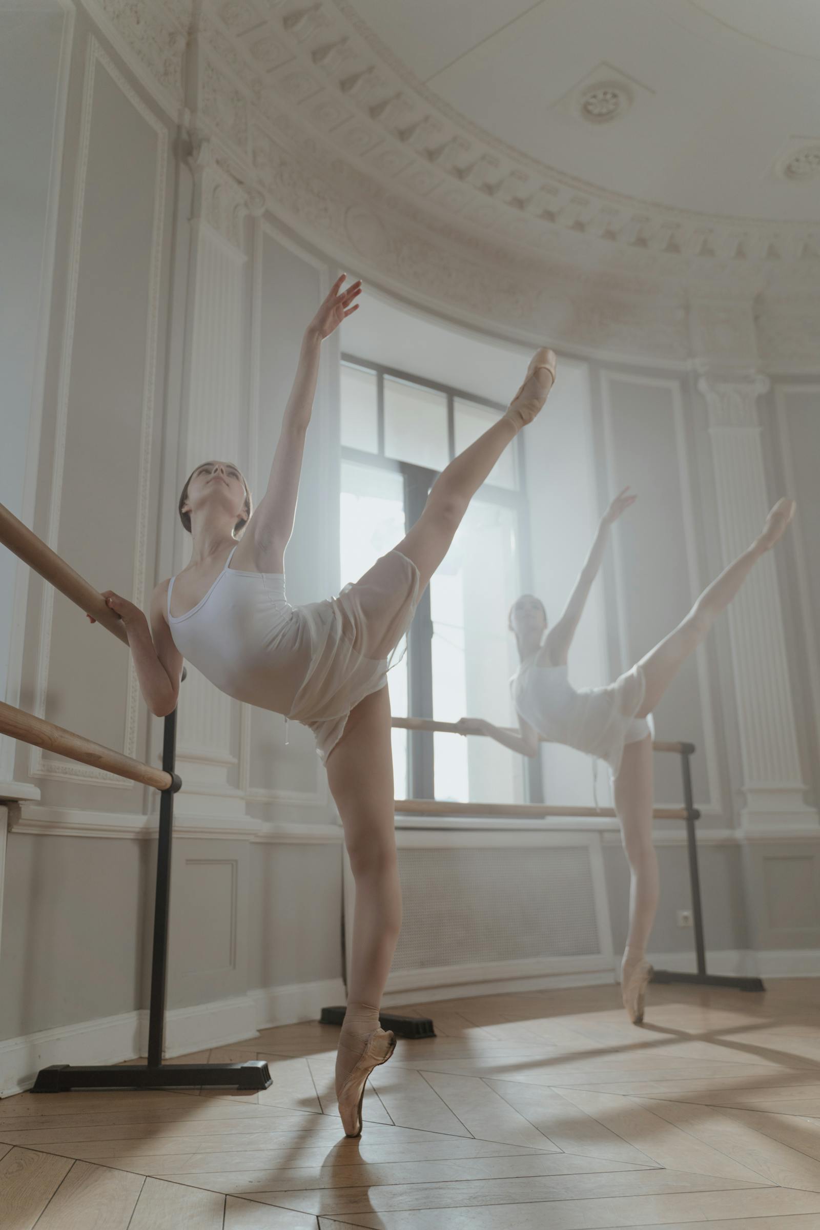 Two dancers in arabesque at the barre in a classical baroque studio — Vaganova-style technical line